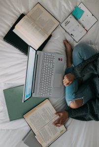 Person sitting on bed with a laptop and open text books representing a student studying